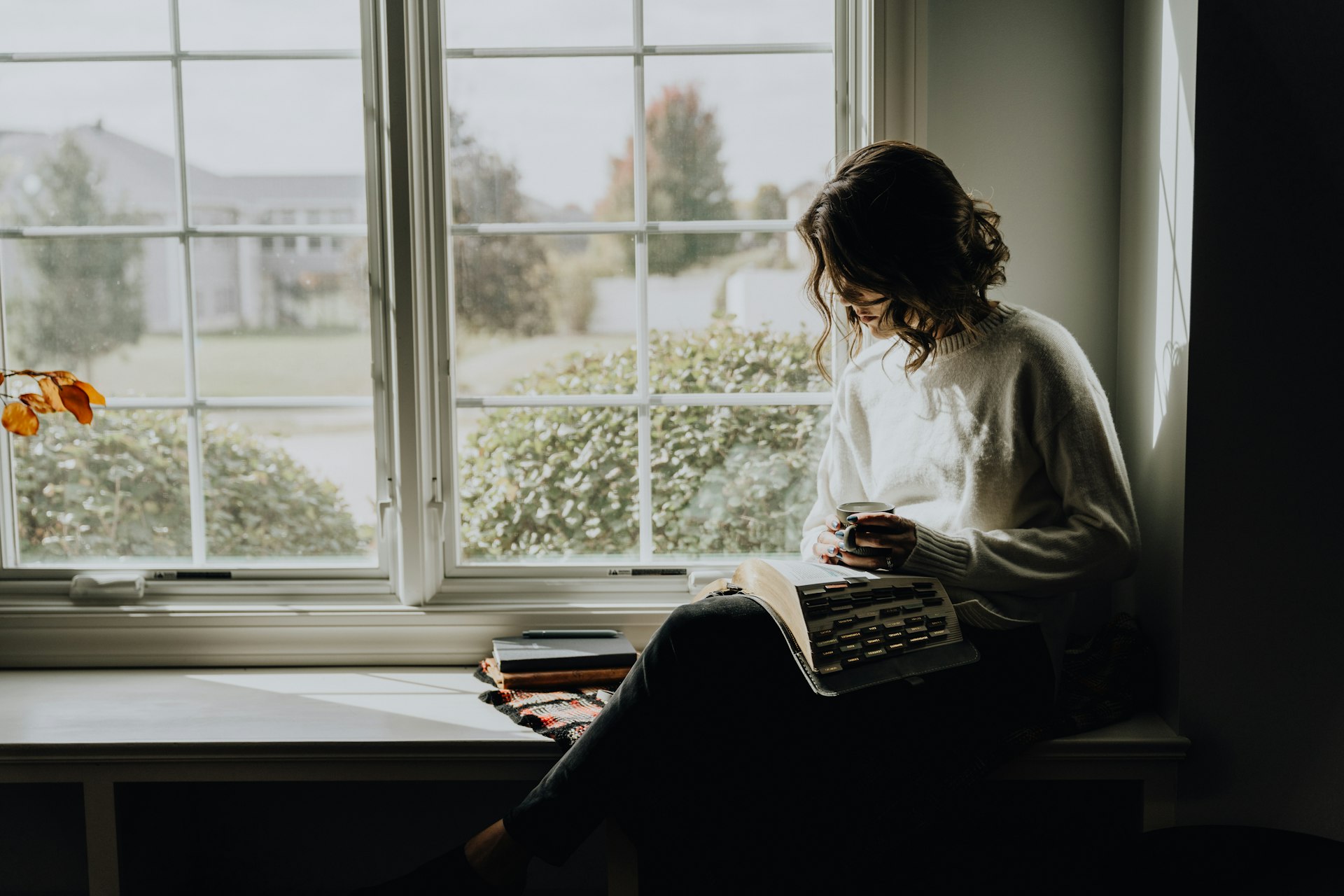 Une femme lit un livre, assise près d'une fenêtre, une tasse à la main